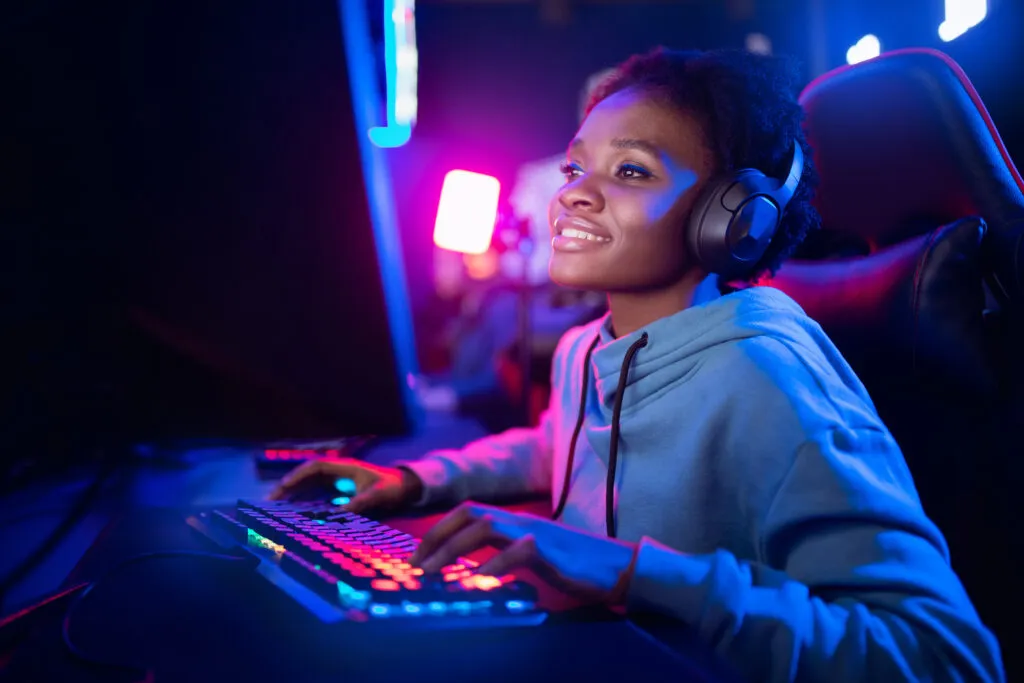 A young, Black, professional woman steams a game on her computer. Her game room has a personal computer, armchair, and a keyboard in neon color.