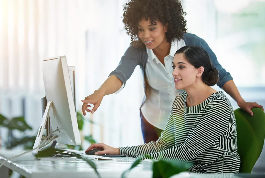 A Black, female presenting mentor stands beside a young intern at her desk. The mentor is pointing to the computer showing the intern how business is done.